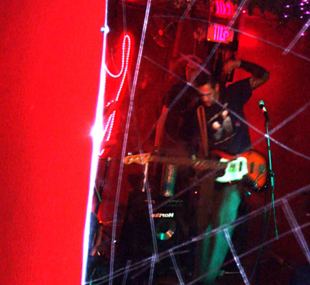 Ein Mann in einem schwarzen T-Shirt steht in der Mitte und hält eine Gitarre, während ein anderer Mann hinter ihm möglicherweise Instrumente spielt, alles unter Scheinwerfern mit einer roten Wand im Hintergrund.