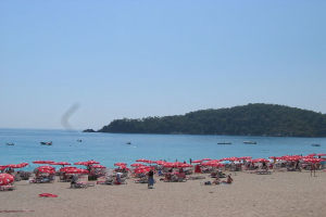 Eine Gruppe von Menschen, die auf Bänken unter Schirmen sitzen und an einem Ufer ruhen, mit Booten auf dem Wasser und Bäumen im Hintergrund bei einem klaren Himmel.