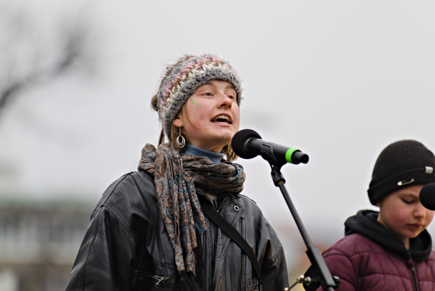 Eine Frau in einer schwarzen Jacke, einem Schal und einer Mütze singt in ein Mikrofon auf der Bühne, während eine andere Person in einer maronenfarbenen Jacke und einer schwarzen Mütze neben ihr steht, vor einem unscharfen Publikum.