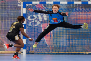 Zwei Frauen beim Handballspielen auf einem Platz mit einem Tor und einer Fahne im Hintergrund, beide tragen Sportschuhe.