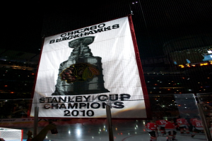 Eine nächtliche Stadionszene mit einem großen Banner in der Mitte, Zuschauern im Hintergrund, einem Glas auf dem Boden und ein paar Spielern auf dem Feld, beleuchtet von Stadionlampen.