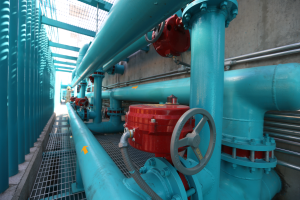 Eine große Gruppe von blauen Rohren und Ventilen in einem Gebäude, mit einem prominenten roten Ventil in der Mitte, Metallstangen auf der linken Seite und einer Wand im Hintergrund, die wahrscheinlich den Bau einer Gasleitung darstellt.