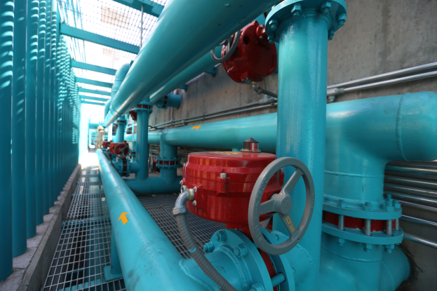 Eine große Gruppe von blauen Rohren und Ventilen in einem Gebäude, mit einem prominenten roten Ventil in der Mitte, Metallstangen auf der linken Seite und einer Wand im Hintergrund, die wahrscheinlich den Bau einer Gasleitung darstellt.