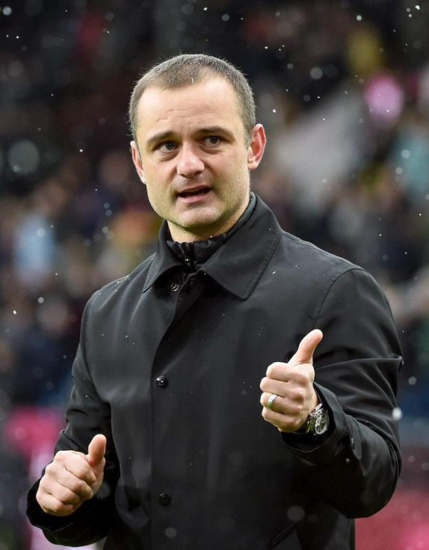 Mann in schwarzer Jacke zeigt Daumen hoch, trägt Uhr, mit unscharfem Hintergrund.