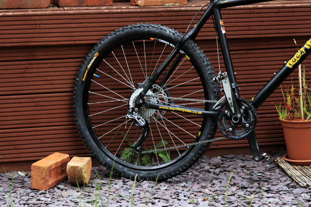 Ein Fahrrad lehnt an einer Wand mit Ziegeln, Steinen und Pflanzen in der Nähe.