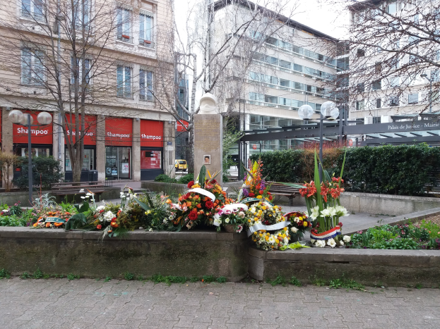 Ein Denkmal in einem Stadtplatz, umgeben von Gebäuden, Bäumen, Pflanzen, Laternen, Bänken und einer Statue, mit Blumen vor ihm auf dem Boden.