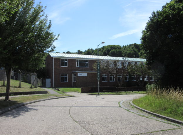Ein Schulgebäude mit Fenstern befindet sich inmitten eines Parks, umgeben von saftigem Grün, mit einem Straßenpfahl, einem Schild, einer Baumgruppe, einem Zaun und einem bewölkten Himmel im Hintergrund.