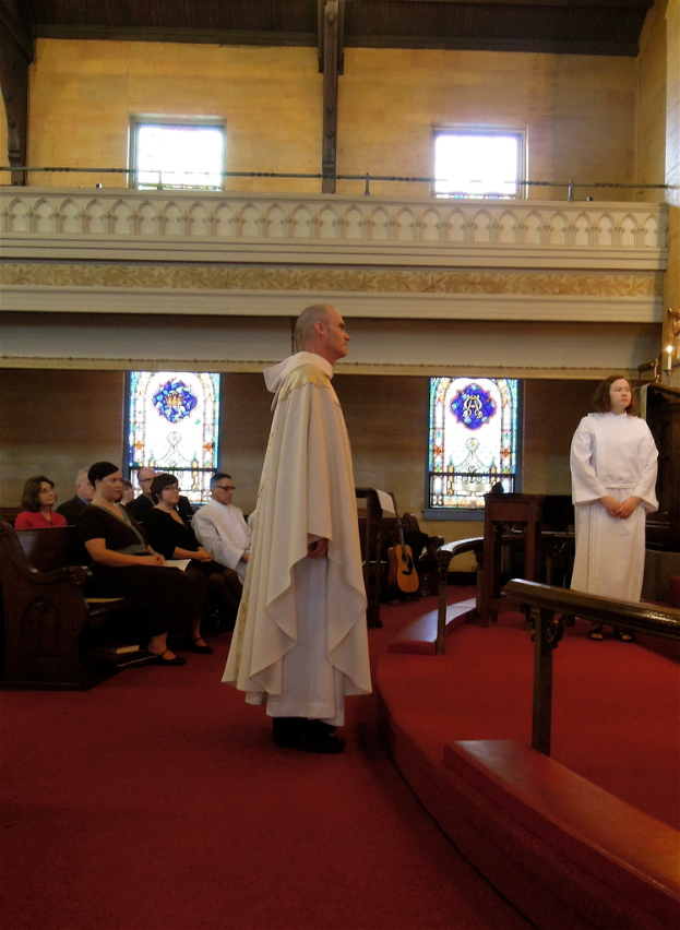 Eine Frau und ein Mann in weißen Kleidern stehen in einer Kirche, mit Menschen auf Bänken links, Musikinstrumenten im Hintergrund und einem roten Teppich, Geländern, Kerzen und Fenstern in der Szene.