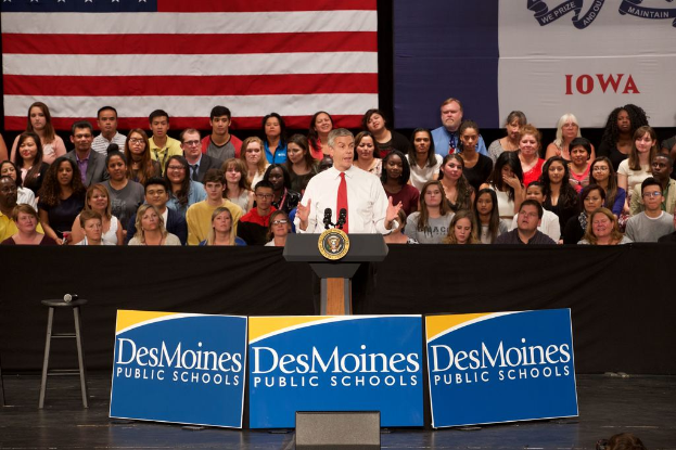 Ein Mann in einem weißen Hemd und roter Krawatte steht an einem Podium mit drei Tafeln, einem Hocker mit Mikrofon und einer sitzenden Menge im Hintergrund, mit einer Flagge und zusätzlichem Text.