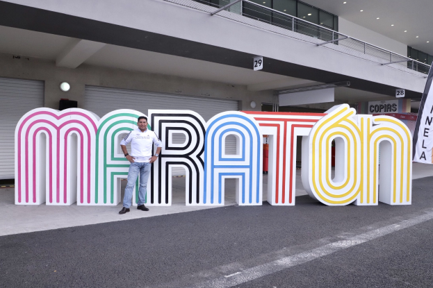 Ein Mann steht am Start eines Marathons vor einem großen "Marathon"-Schild mit einem Banner rechts daneben, im Hintergrund ein Gebäude mit Lichtern, Geländern und Läden.