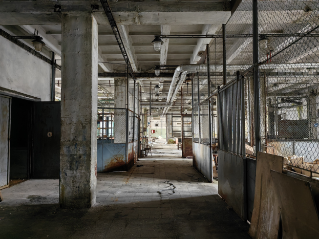 Leerer Raum mit großen Fenstern, Säulen, einer Tür, Deckenleuchten, Metallstangen, einem Maschendrahtzaun, verstreuten Gegenständen auf dem Boden und einer Wand, die eine verlassene Fabrik darstellt.