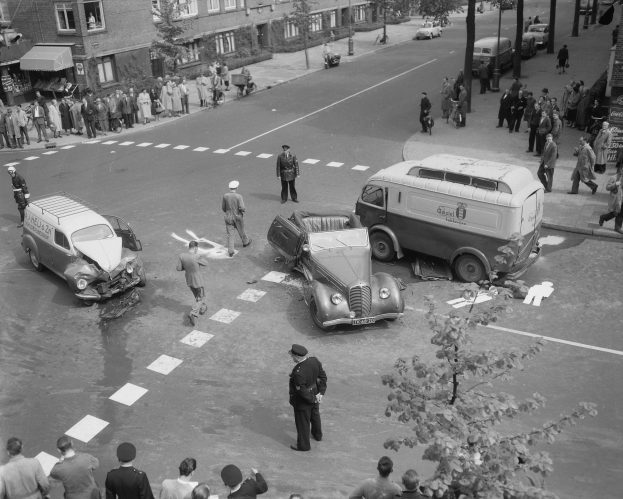 Schwarz-weißes Bild einer Autounfall-Szene an einer Straßenkreuzung mit mehreren Fahrzeugen, darunter ein Lieferwagen, und einer Gruppe von Schaulustigen; Gebäude, Bäume, Pfähle und Texttafeln sind im Hintergrund zu sehen.