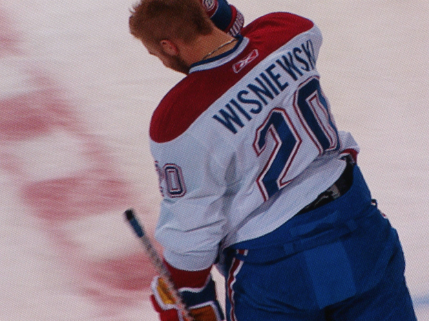 Ein Hockey-Spieler in einem "Wisniewski"-Trikot steht auf dem Eis und hält einen Hockey-Schläger, mit konzentrierter und entschlossener Miene.