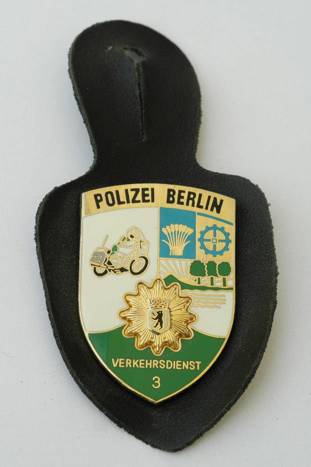 Ein schildförmiges Polizeisiegel mit der Aufschrift "Polizei Berlin" in fetter schwarzer Schrift auf einem blauen Hintergrund mit einem weißen Stern und Rahmen, das auf einer Oberfläche liegt.