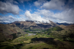 Eine Panoramansicht vom Gipfel eines Berges in Queenstown, Neuseeland, zeigt saftiges grünes Gras, verstreute Bäume und eine gewundene Straße unter einem Himmel voller weißer, flauschiger Wolken.