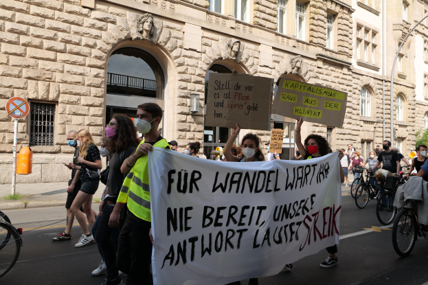 Eine Gruppe von Menschen marschiert auf einer Straße, einige halten Schilder und andere fahren Fahrräder, im Hintergrund ein Gebäude mit Bögen, Säulen und Skulpturen.