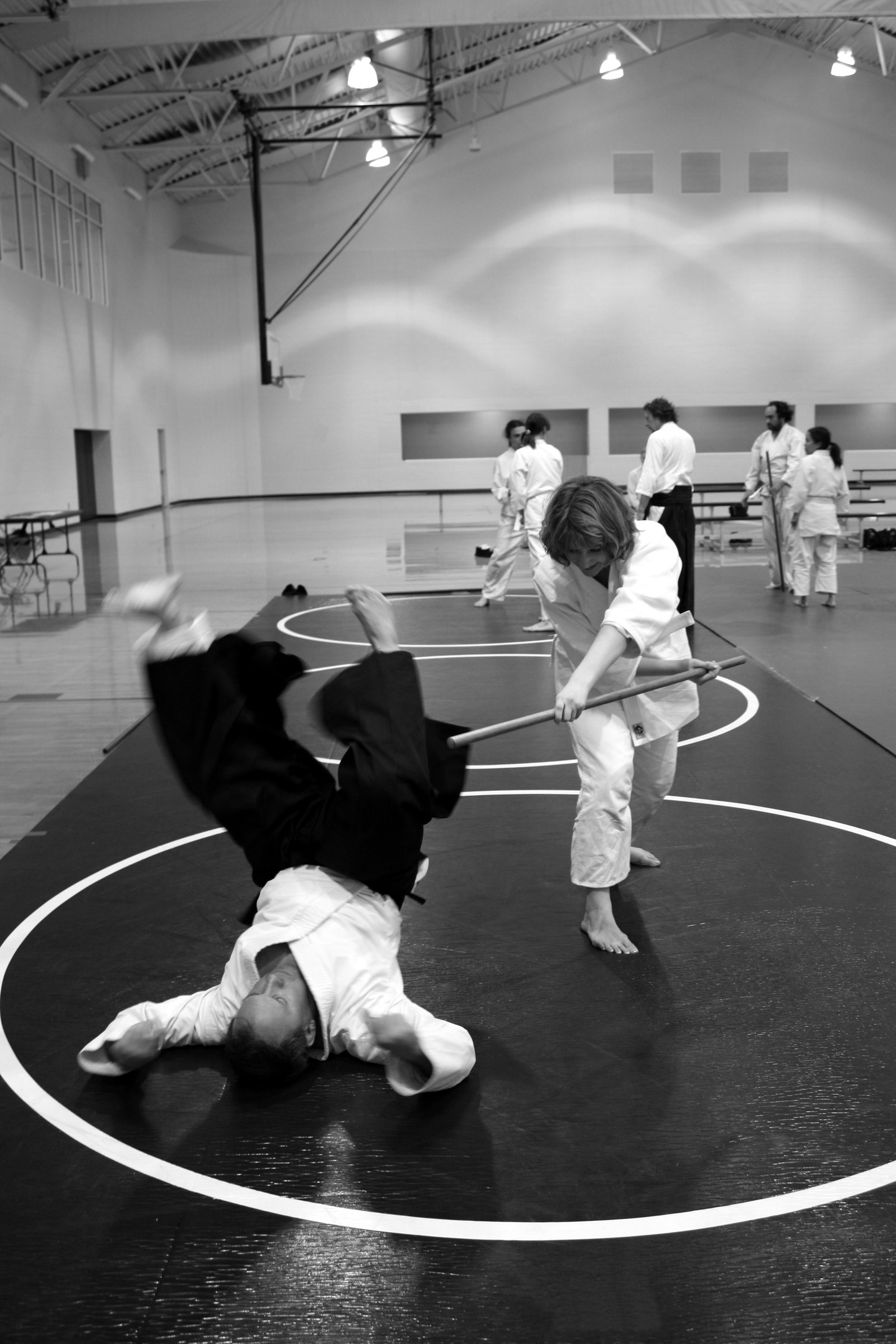 Zwei Personen in einer Karate-Halle üben Stockkampf, während andere zuschauen, mit Leuchten entlang der Wand im Hintergrund.