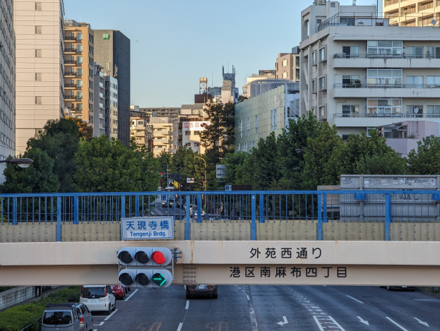 Stadtstraße mit Fahrzeugen, eine Brücke mit Geländern und einem Schild, Verkehrsampeln, Laternenpfähle, Bäume, Gebäude mit Fenstern und ein klarer Himmel.