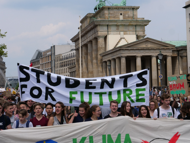 Gruppe von Schülern marschiert in Berlin mit einem bunt bemalten 'Students for Future'-Schild vor einer Kulisse aus Gebäuden, Bäumen und Himmel.