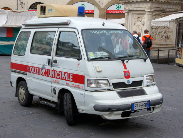 Ein weißer Van fährt auf einer Straße mit einer Gruppe von Menschen in der Nähe, die Taschen tragen, ein Schuppen auf der rechten Seite und Gebäude und Metallstangen im Hintergrund.