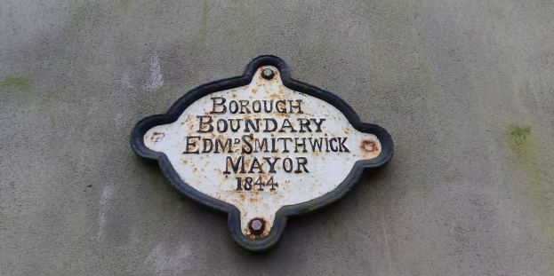 Eine Metalltafel an einem Gebäude mit schwarzem Hintergrund und weißer, fetter, zentrierter Schrift, die 'Borough Boundary Edm Smithwick Mayor' lautet.
