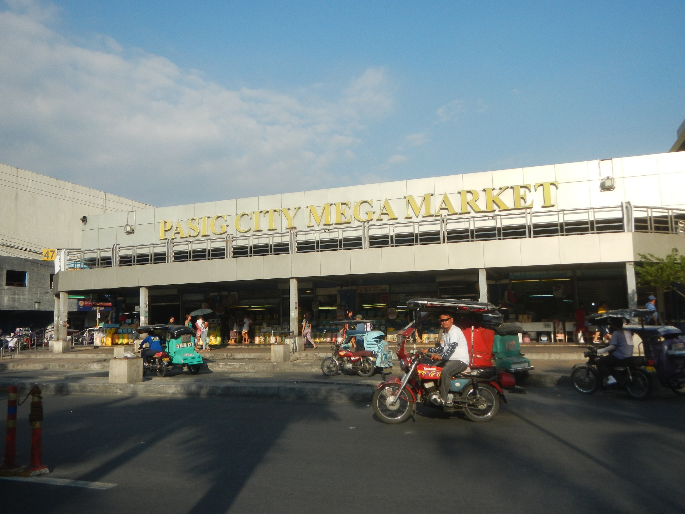 Ein belebter Pasig City Mega Market mit Fahrzeugen auf der Straße und Fußgängern, mit einem Gebäude mit Text im Hintergrund unter einem bewölkten Himmel.