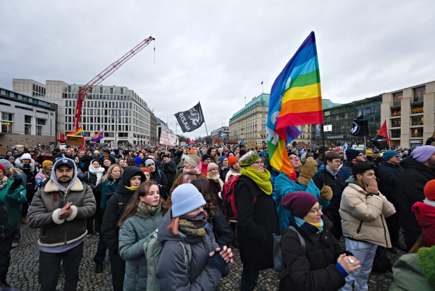 Eine große Gruppe von Menschen bei einer LGBTQ+-Rechtsdemo in Berlin, die Fahnen und Schilder halten, mit Gebäuden und einem Kran im Hintergrund.