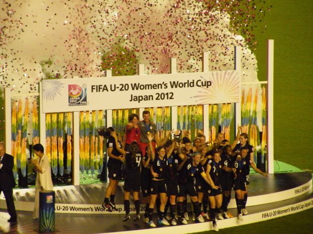 Gruppe von Frauenfußballspielerinnen in schwarzen Uniformen, die auf einer Bühne feiern, umgeben von einer Menge mit Kameras, mit einem dekorierten Hintergrund und grasbewachsenem Boden.