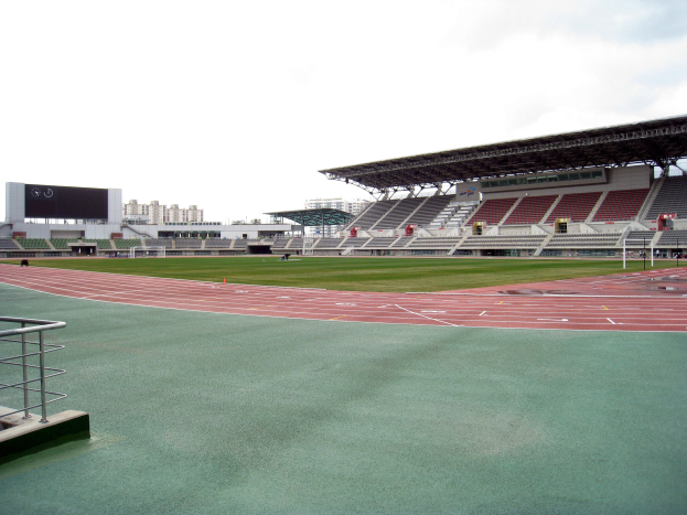 Großes Stadion mit zentraler Laufbahn, einer Begrenzung auf der linken Seite, Gebäuden im Hintergrund und einem klaren blauen Himmel.