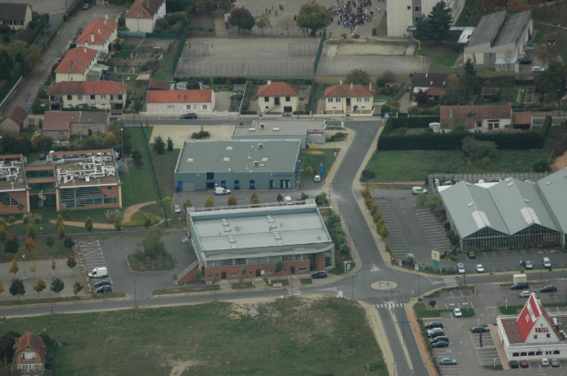 Eine Vogelperspektive auf ein dicht bebautes Stadtgebiet mit zahlreichen Gebäuden, Straßen voller Fahrzeuge, geparkten Autos und zwischendurch Bäumen und Gärten.