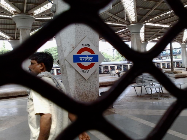 Ein Mann steht hinter einem Gitter auf einer Plattform, mit Bänken und Säulen in der Nähe, unter einem Dach, mit einem Zug, einer Brücke und Bäumen im Hintergrund.