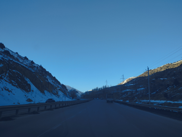 Ein Auto fährt auf einer schneebedeckten Straße mit Geländern und Strommasten, mit schneebedeckten Bergen im Hintergrund und einem klaren Himmel darüber.