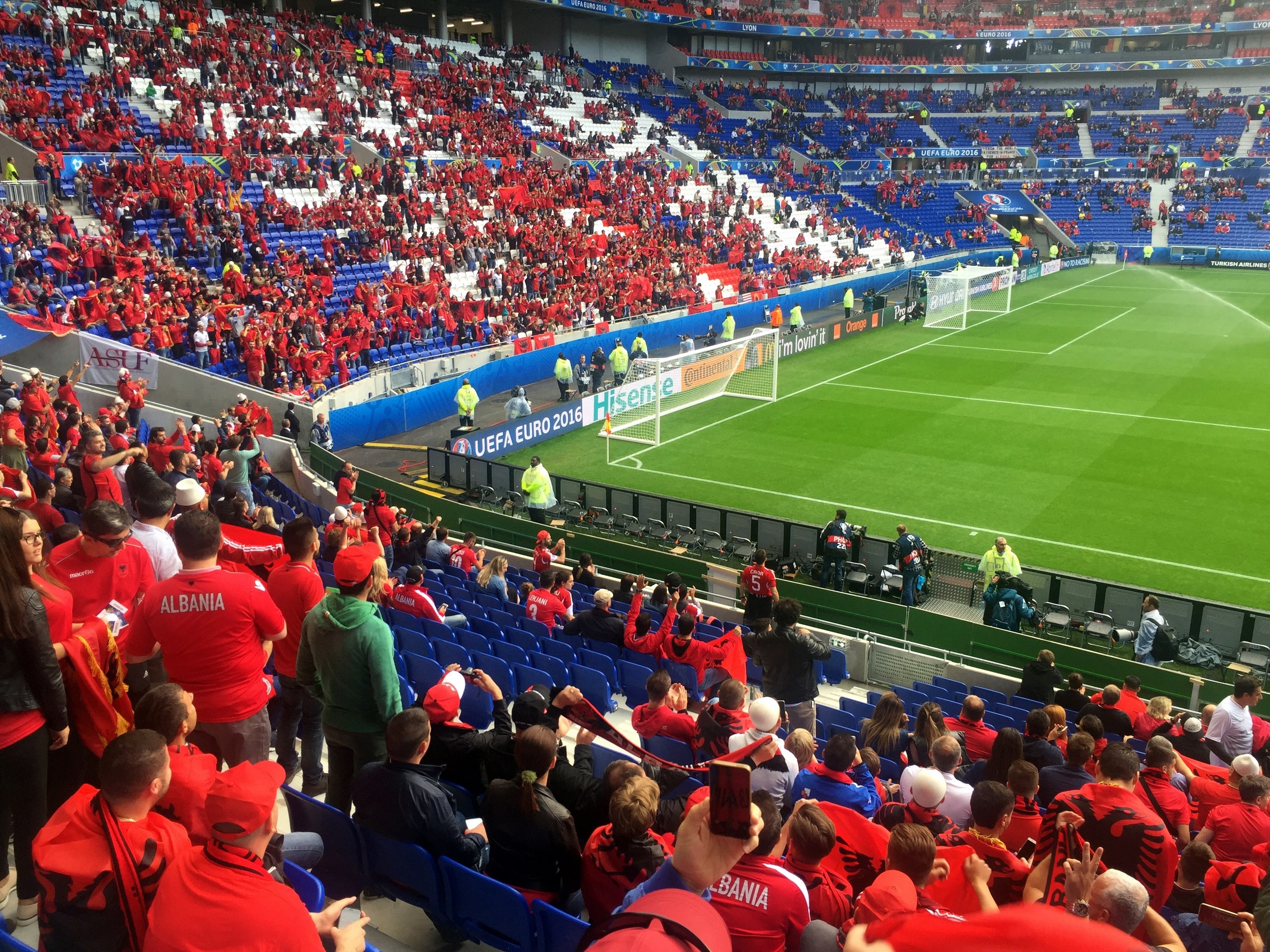 Eine große Menschenmenge schaut einem Fussballspiel zu, mit Menschen auf Sitzen und Stehplätzen, Bannern, einem Torpfosten und Deckenleuchten.
