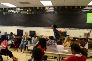 Ein Klassenzimmer mit Kindern an Tischen mit verschiedenen Gegenständen, einer Lehrerin in der Mitte und einer Tafel im Hintergrund.