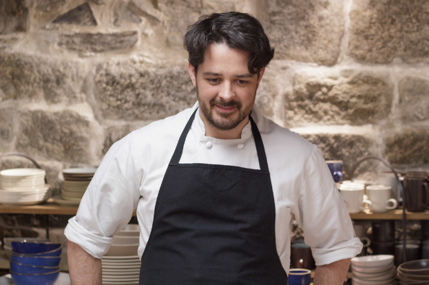 Ein Koch in einem weißen Hemd und schwarzer Schürze steht vor einem Regal mit Tellern, Schüsseln und Tassen an einer Wand.