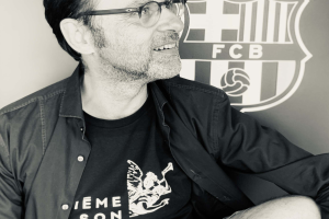 Ein Mann mit Brille und schwarzem Hemd lächelt, während er vor dem FC Barcelona-Logo an einer Wand sitzt, auf einem Schwarz-Weiß-Foto.