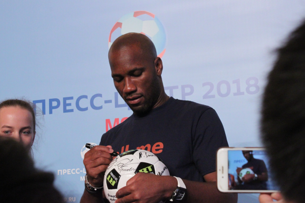 Ein Mann signiert einen Fußball mit einem Stift, während er ihn hält, und eine Frau steht neben ihm; eine Person fotografiert die Szene mit einem Handy, und ein Banner ist im Hintergrund sichtbar.