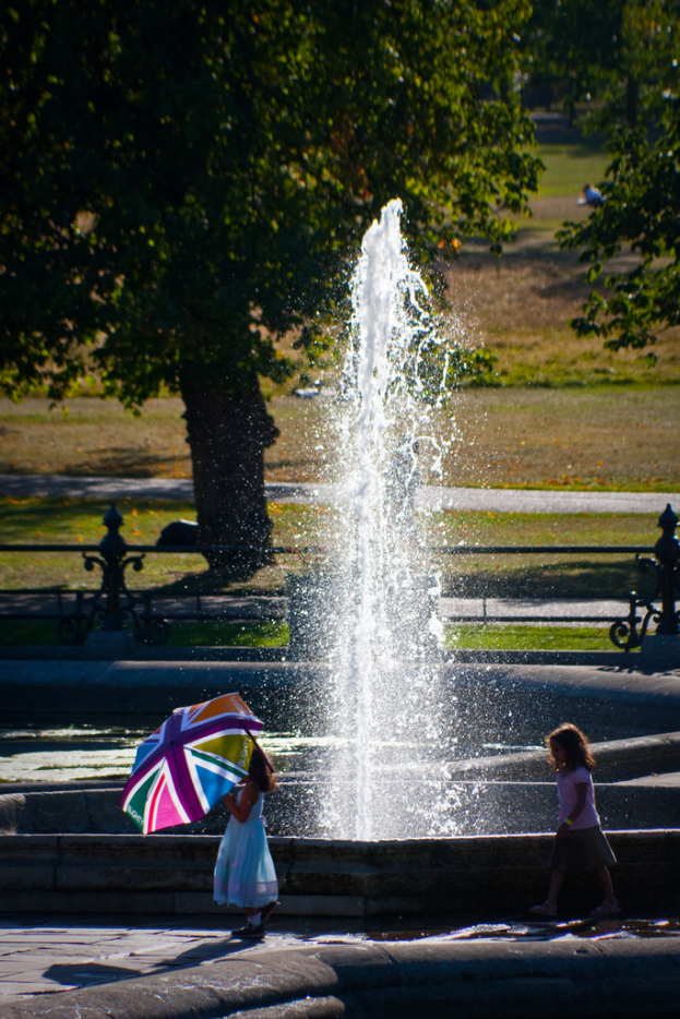 Zwei Mädchen vor einem Brunnen, eine hält einen Schirm, mit Bäumen und Wiese im Hintergrund.