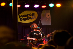 Eine Person in einem schwarzen Kleid singt und spielt Gitarre auf der Bühne, mit einem Mikrofon, Trommeln, einem blauen Vorhang mit einem Plakat und Bühnenlichtern dahinter und einer Menge davor.