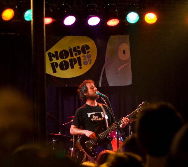 Eine Person in einem schwarzen Kleid singt und spielt Gitarre auf der Bühne, mit einem Mikrofon, Trommeln, einem blauen Vorhang mit einem Plakat und Bühnenlichtern dahinter und einer Menge davor.