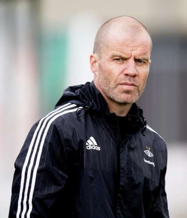 Glatzköpfiger Mann in einem blauen Jackett mit weißen Streifen und Kragen, der auf einem Fussballfeld steht und die Hände verschränkt hat.