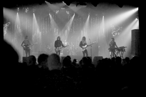 Ein Schwarz-Weiß-Bild eines Musikkonzerts mit vier Künstlern auf der Bühne, darunter drei Gitarristen und eine Sängerin, sowie Zuschauern unten und Scheinwerfern oben.