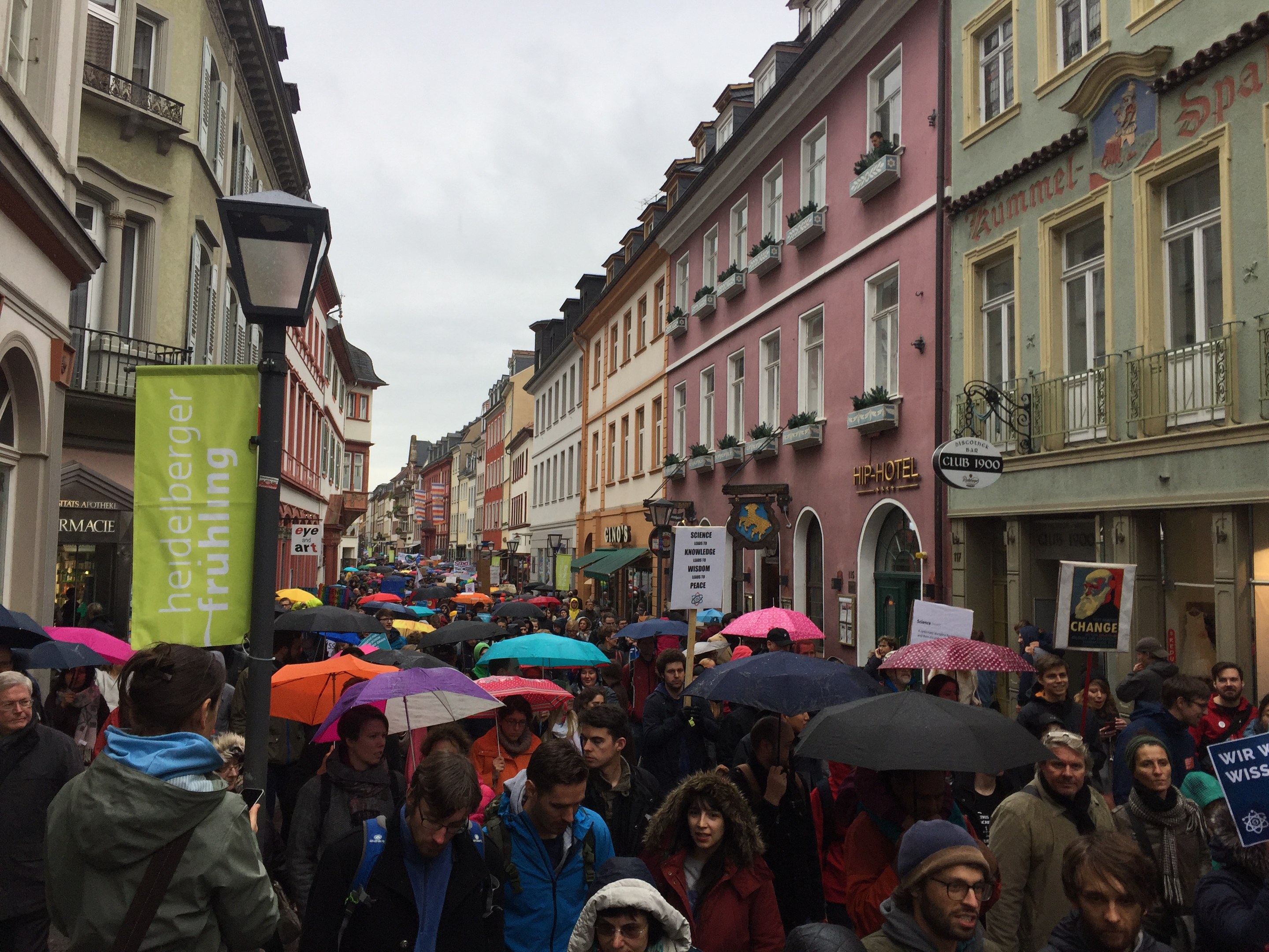 Große Gruppe von Menschen, die mit Schirmen die Straße entlanggehen, einige tragen Taschen und halten Schilder, mit Gebäuden auf beiden Seiten und einem Laternenmast im Vordergrund.