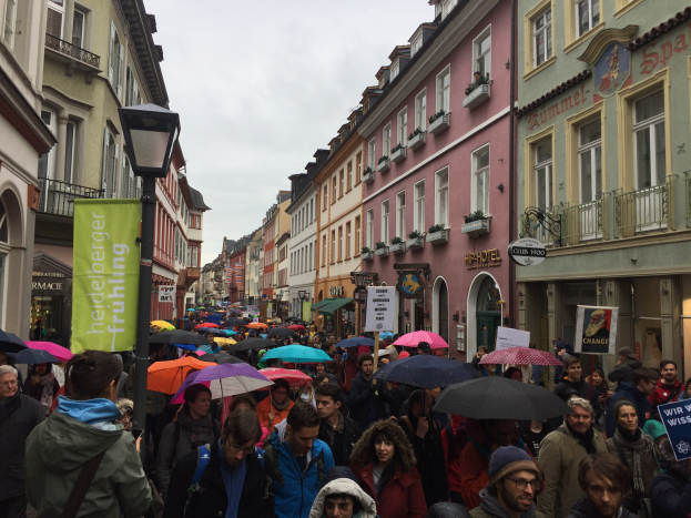 Große Gruppe von Menschen, die mit Schirmen die Straße entlanggehen, einige tragen Taschen und halten Schilder, mit Gebäuden auf beiden Seiten und einem Laternenmast im Vordergrund.