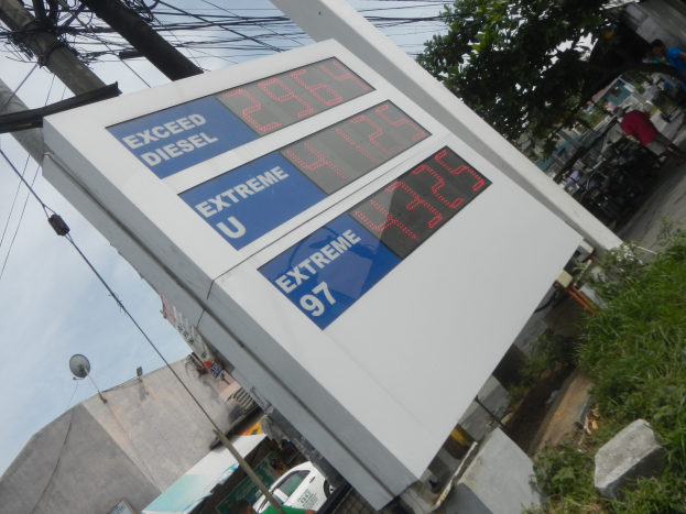 Eine Tankstelle mit einem Schild, auf dem "Exceed Diesel" steht, im Vordergrund mit Strommästen, Gebäuden, Fahrzeugen, Menschen, Bäumen und anderen Gegenständen im Hintergrund unter einem sichtbaren Himmel.