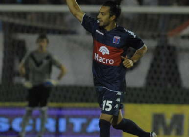 Ein Fußballspieler feiert auf dem Boden nach einem Tor, mit jubelnden Zuschauern und einer Person im Hintergrund, alles vor einem unscharfen, energischen Hintergrund.