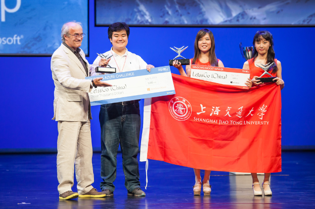 Vier Personen stehen auf einer Bühne, jede hält Auszeichnungen, Schecks und eine Fahne, sie lächeln mit einem digitalen Bildschirm im Hintergrund.