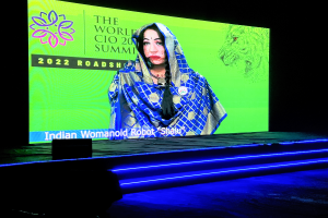 Eine Frau in einem blauen und weißen Kleid steht auf einer Bühne bei der World CIO Summit 2022, beleuchtet von hellen Scheinwerfern, mit einem dunklen Hintergrund und einem großen Bildschirm dahinter.