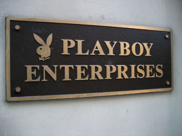 Ein rechteckiger Namensschild mit Logo und Buchstaben, an einer Wand mit sichtbaren Schrauben befestigt.