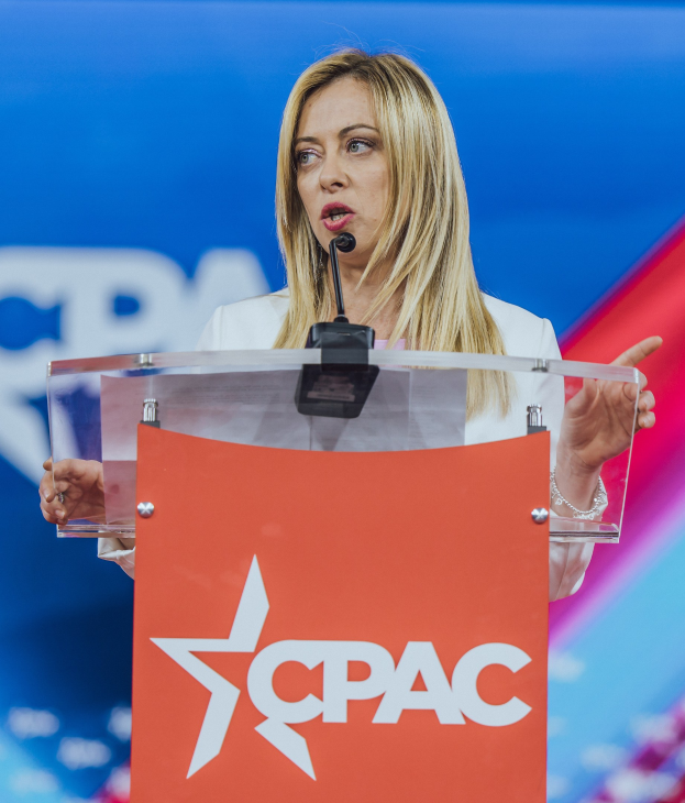 Frau in weißem Kleid, die an einem Rednerpult mit Mikrofon spricht, wahrscheinlich eine Politologin, mit unscharfem Hintergrund.